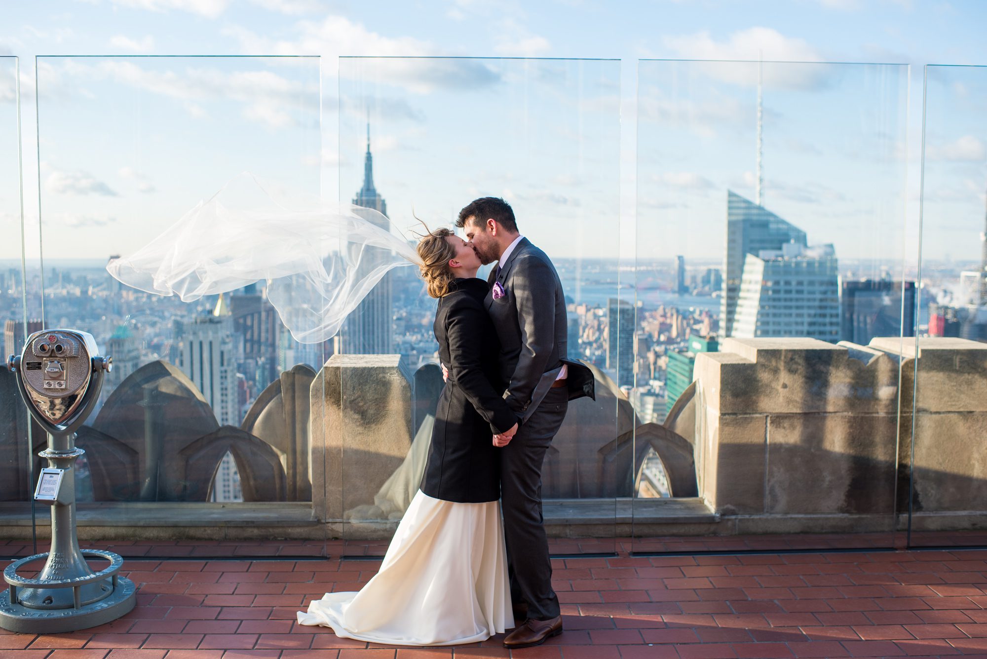 A bride with her veil blowing in the wind kisses her new husband after they eloped at Top of the Rock in NYC. 