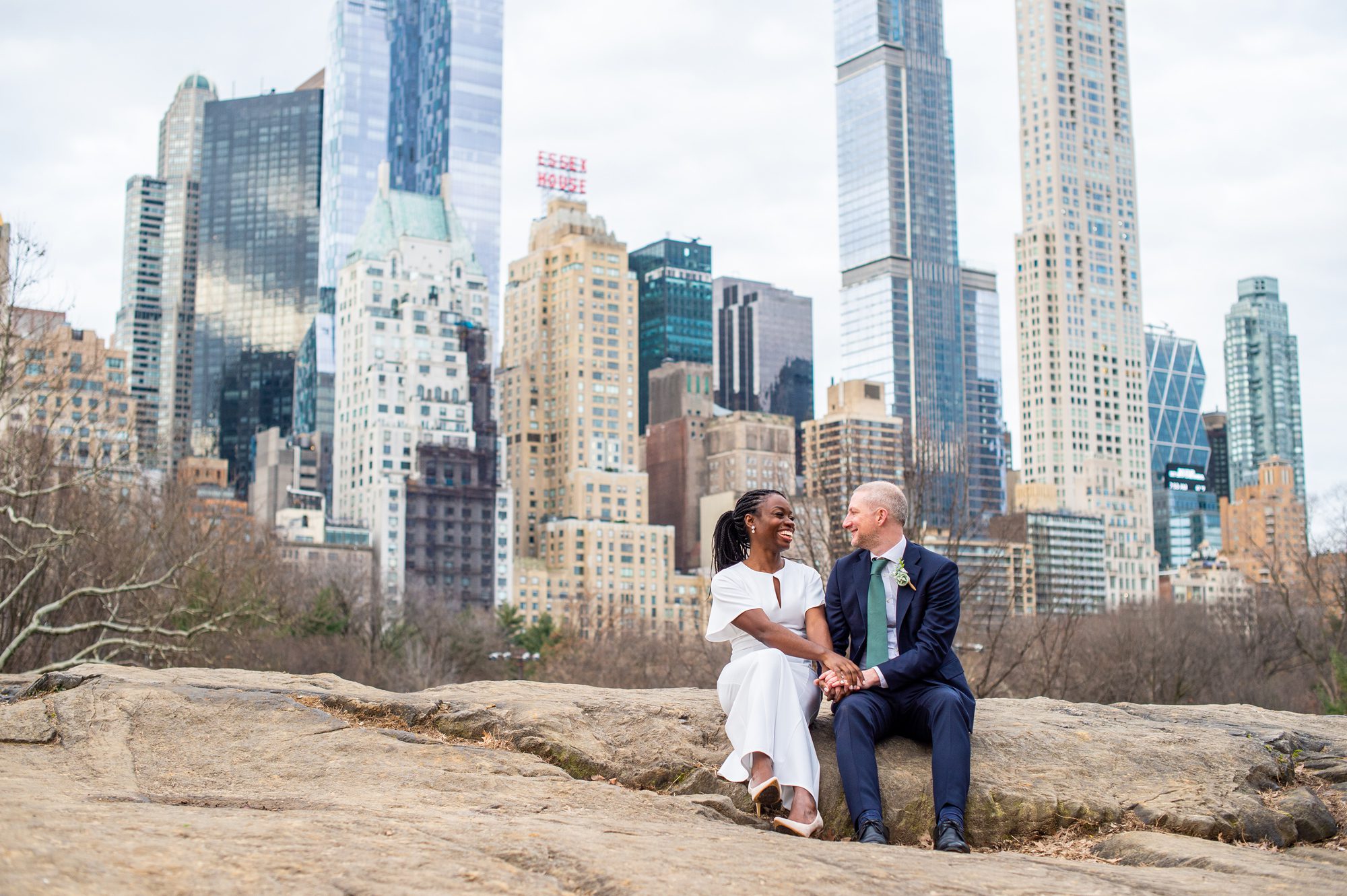 A couple sitting on a rock with the NYC skyline behind them after their Central Park wedding