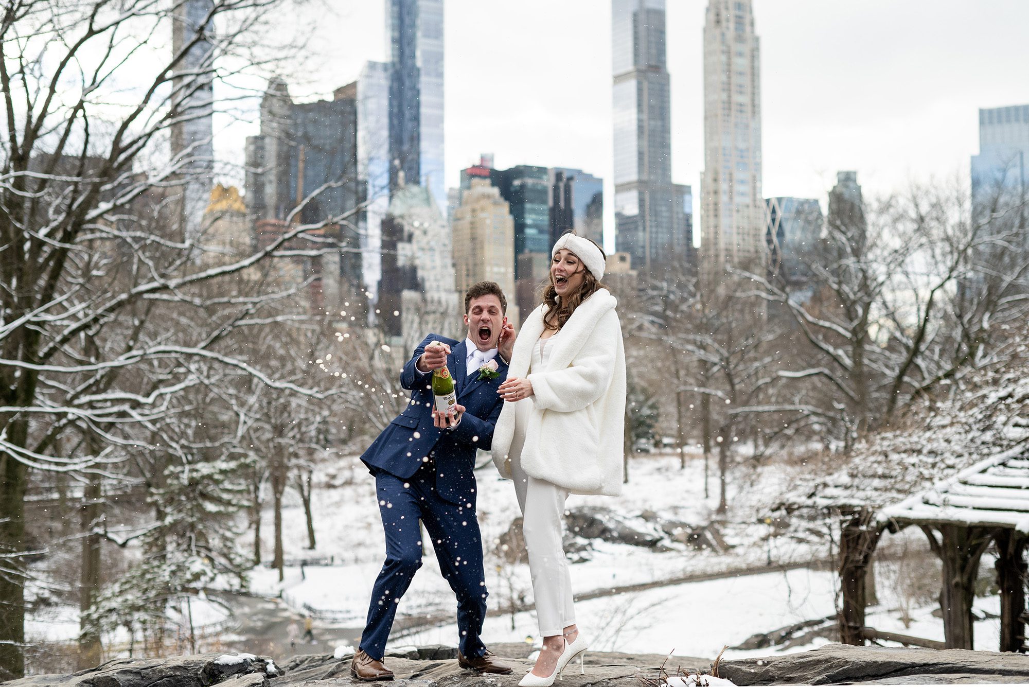 Wedding couple popping a bottle of champagne after their NYC Elopement overlooking the NYC skyline in winter time.