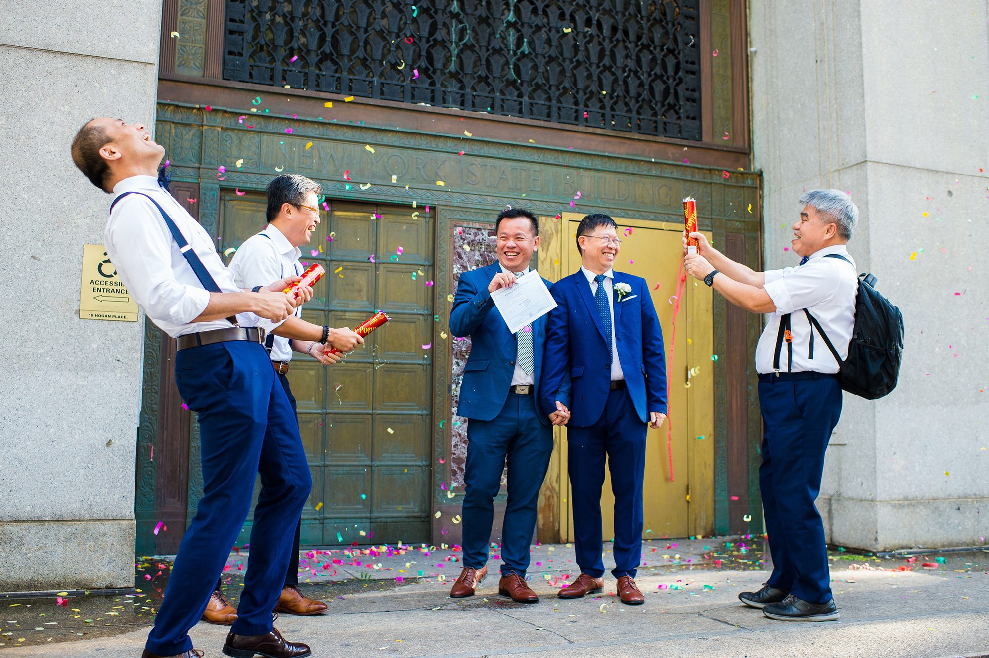 Two grooms exiting City Hall with confetti poppers. 