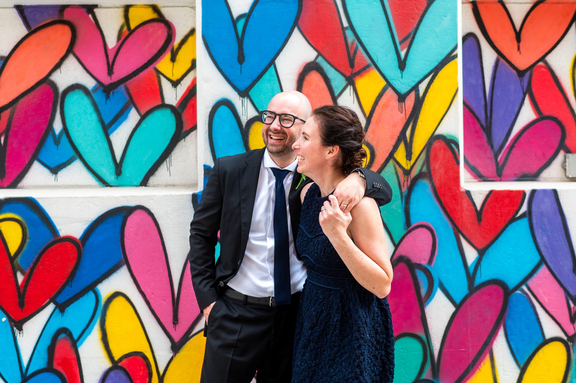A couple laughing in front of a rainbow heart wall in Dumbo Brooklyn