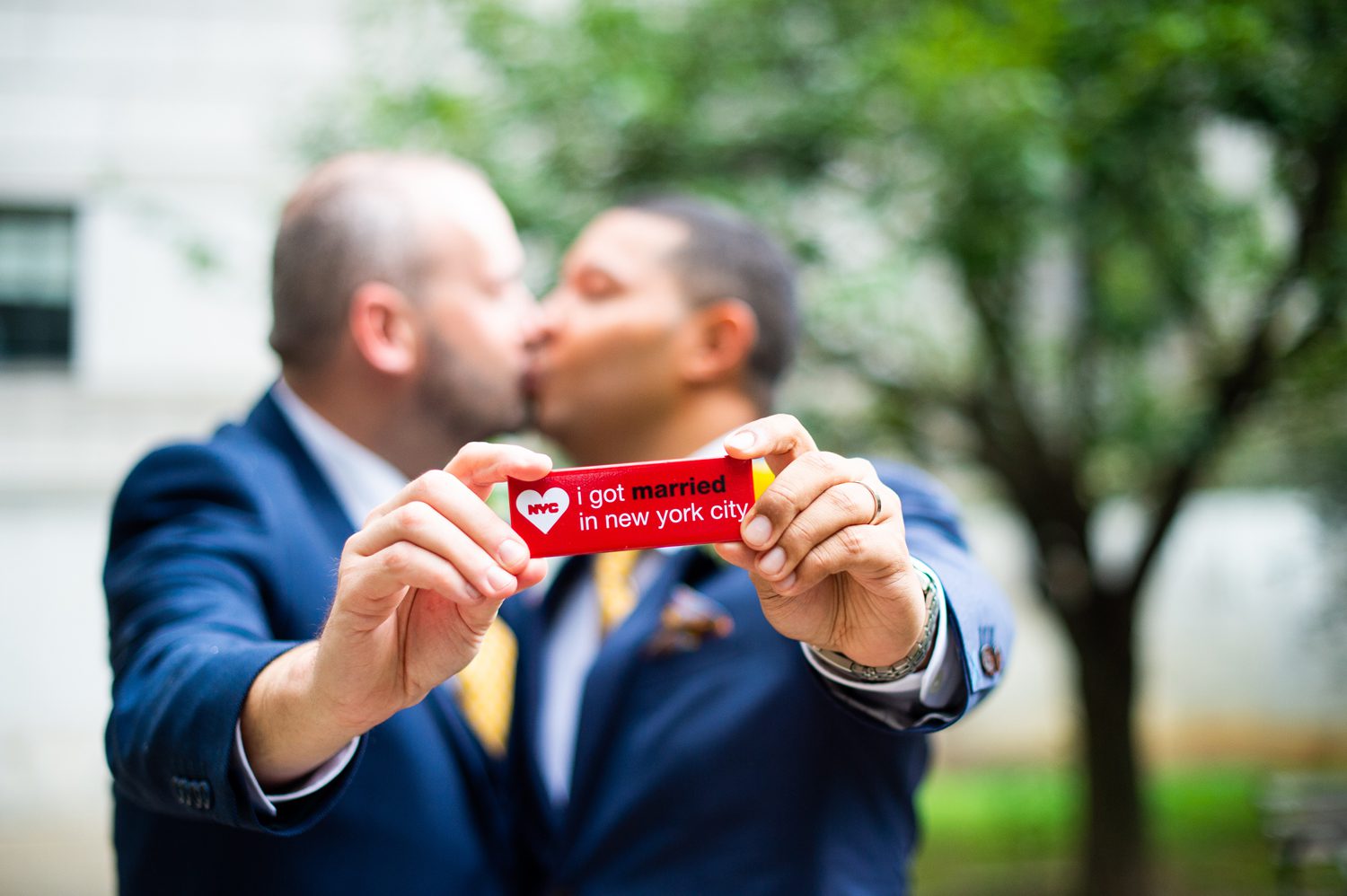 Two grooms kissing holding up a magnet that says we got married in NYC. 