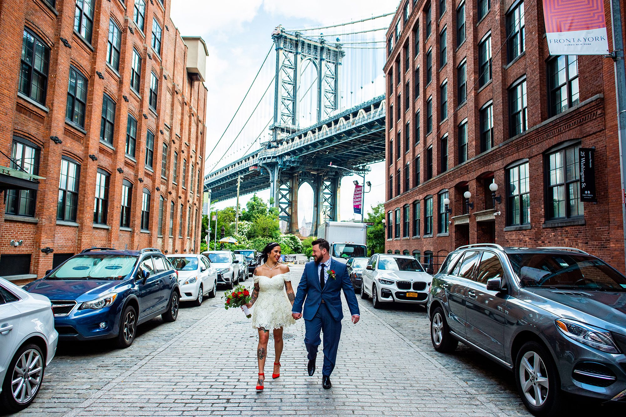 Bride and Groom walk down Washington Street in Dumbo Brooklyn with the Manhattan Bridge behind them. The bride is wearing a short dress, bright red heels and has lots of tattoos. 