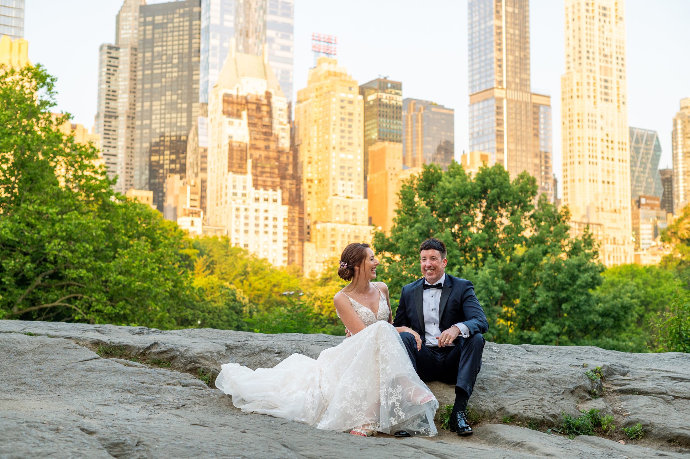 Bride and groom sitting on a rock with NYC skyline behind them at sunrise in Central Park 